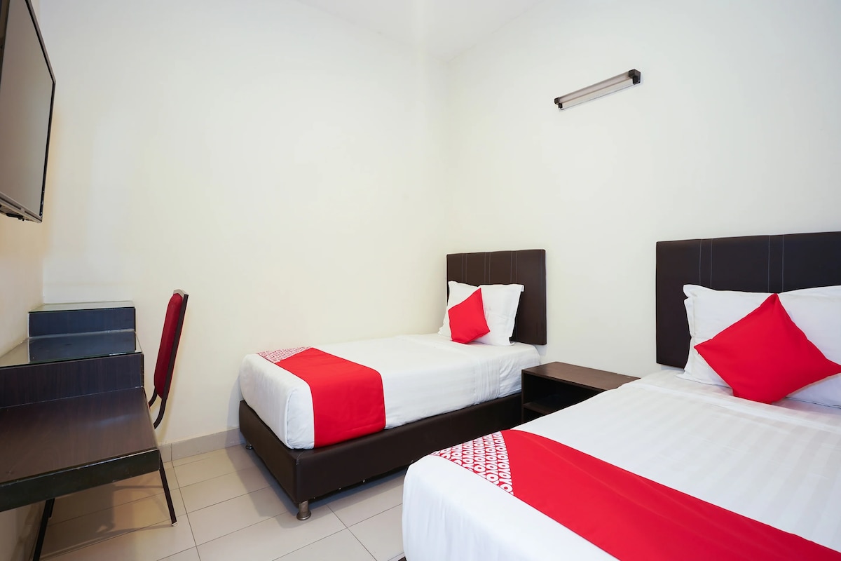 A clean and modern guest room is presented with two spacious beds, each adorned with red accent pillows. A small desk and chair are positioned against the wall, alongside a flat-screen TV mounted above. The bright, light-coloured walls enhance the room's openness.