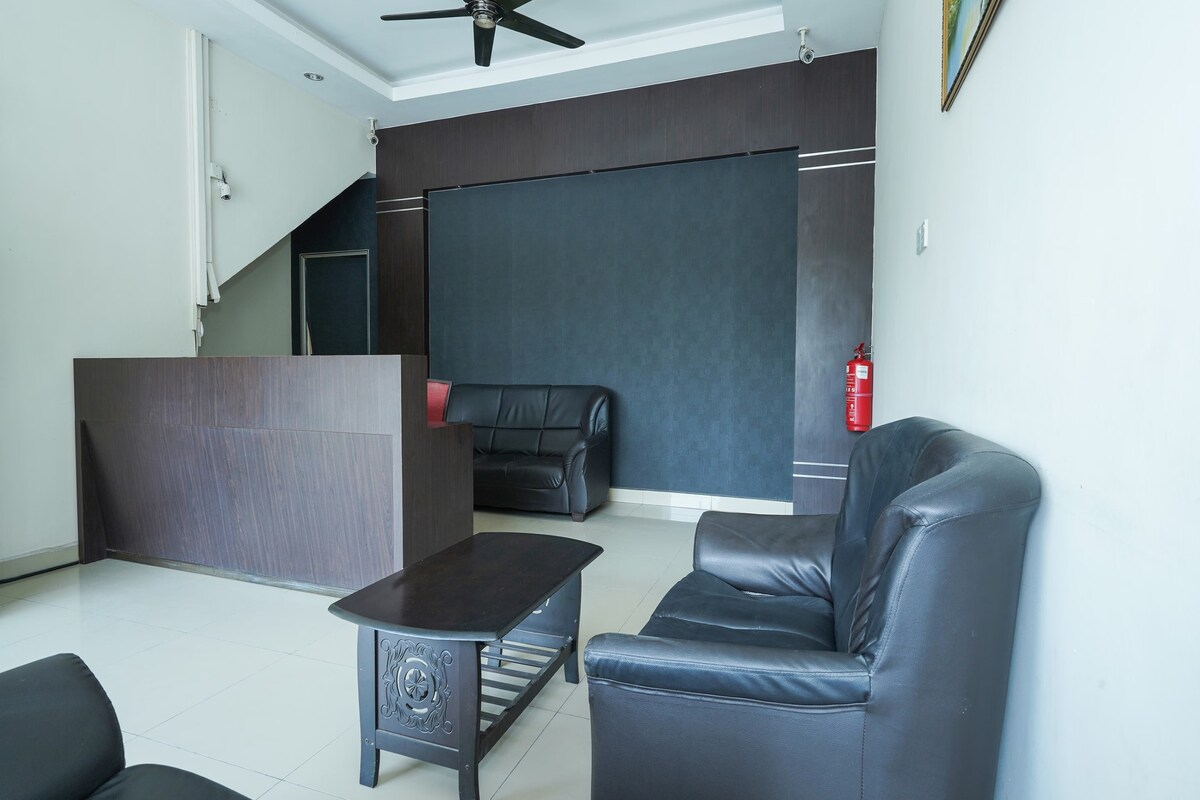 A welcoming lobby area is presented with a sleek reception desk and two black leather sofas arranged around a wooden coffee table. The clean, tiled floor and neutral wall colors create a bright and open atmosphere, complemented by a ceiling fan above.
