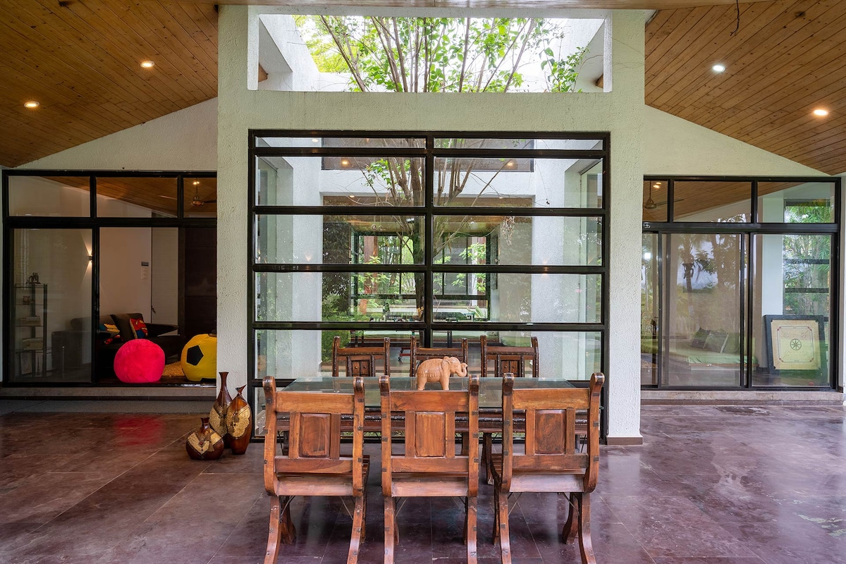 A spacious seating area features a wooden table surrounded by four chairs, positioned beneath a ceiling with wooden accents. Large glass doors provide views of the outdoor greenery, enhancing the connection with nature. Colorful seating options are visible in the background.