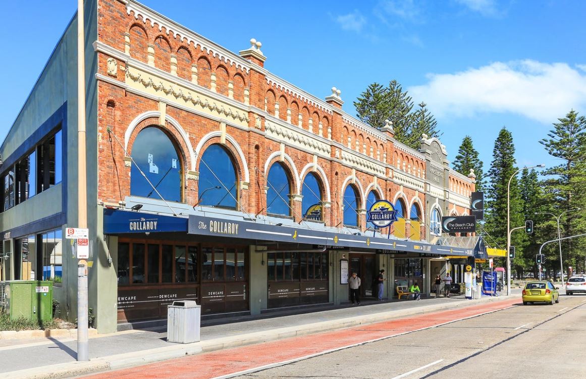A red-brick building features arches and intricate architectural details, housing various shops and eateries. Large windows allow natural light to illuminate the interior. Trees and a blue sky are visible in the background, creating a lively street atmosphere.