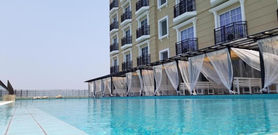 An infinity swimming pool reflects the clear sky, surrounded by elegantly draped cabanas. The pool area features a modern design, with smooth tiles extending towards the horizon, offering serene views of the outdoors and inviting relaxation.