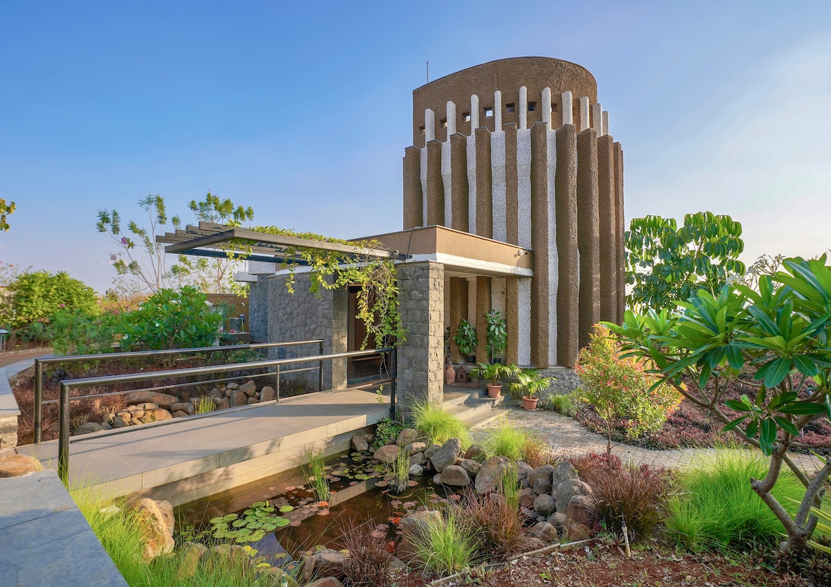 The villa showcases an architectural design featuring a unique circular structure with vertical columns. A stone pathway leads to the entrance, flanked by greenery and a tranquil pond, creating a serene outdoor environment that invites exploration.