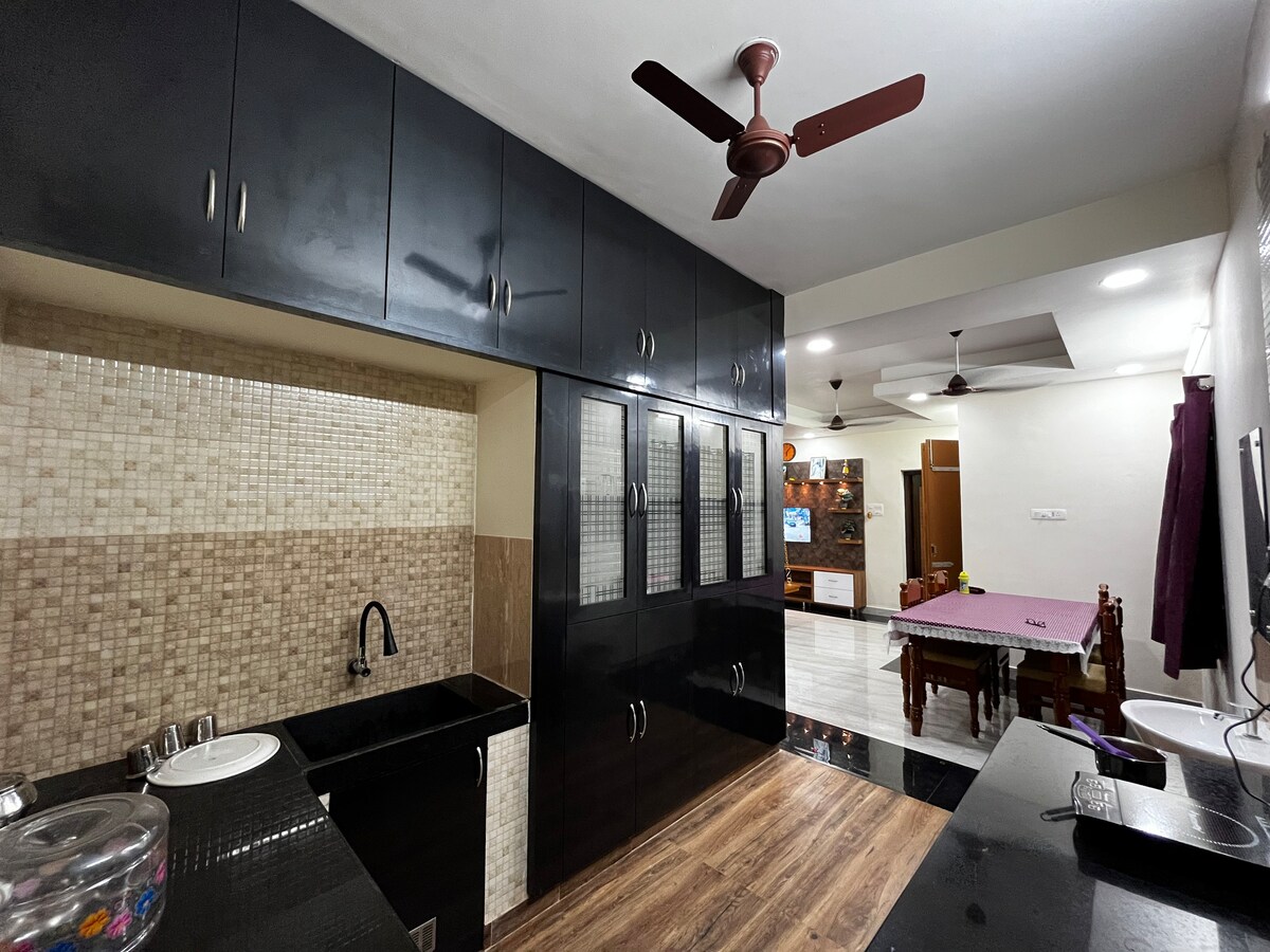 A modern kitchen space is showcased, featuring sleek black cabinetry and a patterned backsplash. A sink is situated next to a countertop, and cabinets with glass panels provide additional storage. The adjacent dining area includes a spacious table and chairs, illuminated by ceiling fans.
