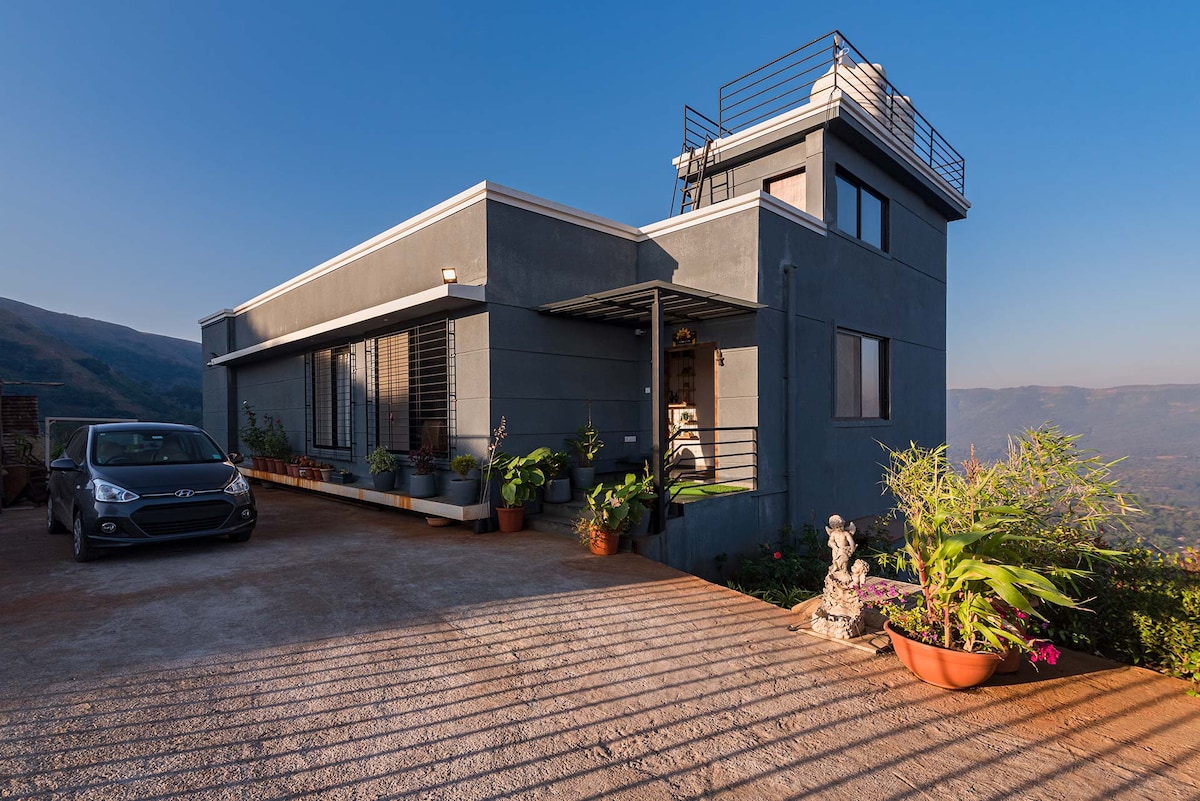 A modern villa is showcased with a sleek concrete exterior, complemented by large windows allowing natural light. Potted plants line the entrance, adding a touch of greenery. A parked car and a stone feature are visible on the well-maintained driveway, enhancing the welcoming ambiance.