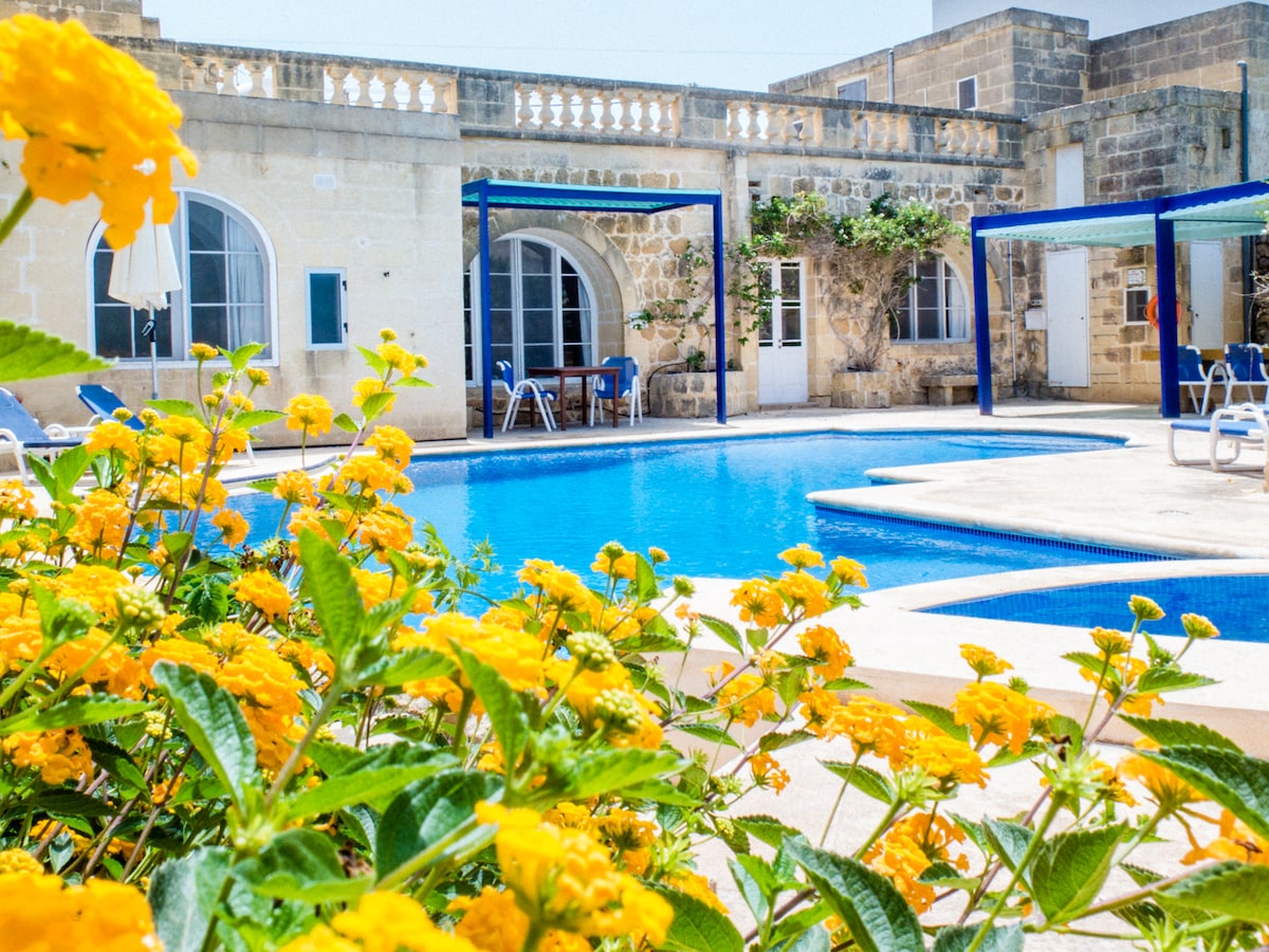 A large swimming pool is surrounded by sunny flower beds, featuring vibrant yellow blooms. The pool area is equipped with shaded seating under blue pergolas, and the original stone walls create a rustic backdrop. Several lounge chairs are visible near the water.