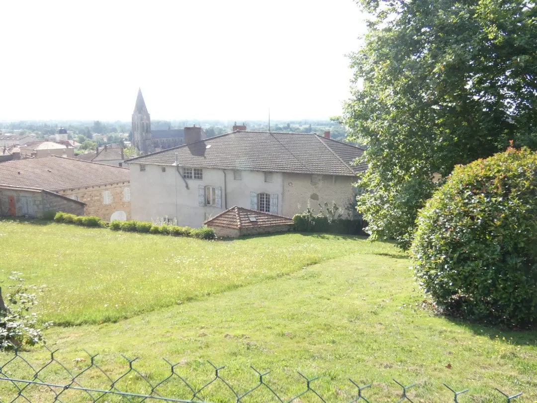 A serene view captures a charming house surrounded by a lush green lawn, framed by neatly trimmed bushes. In the background, a church steeple is visible, towering over the picturesque landscape under bright, clear skies.