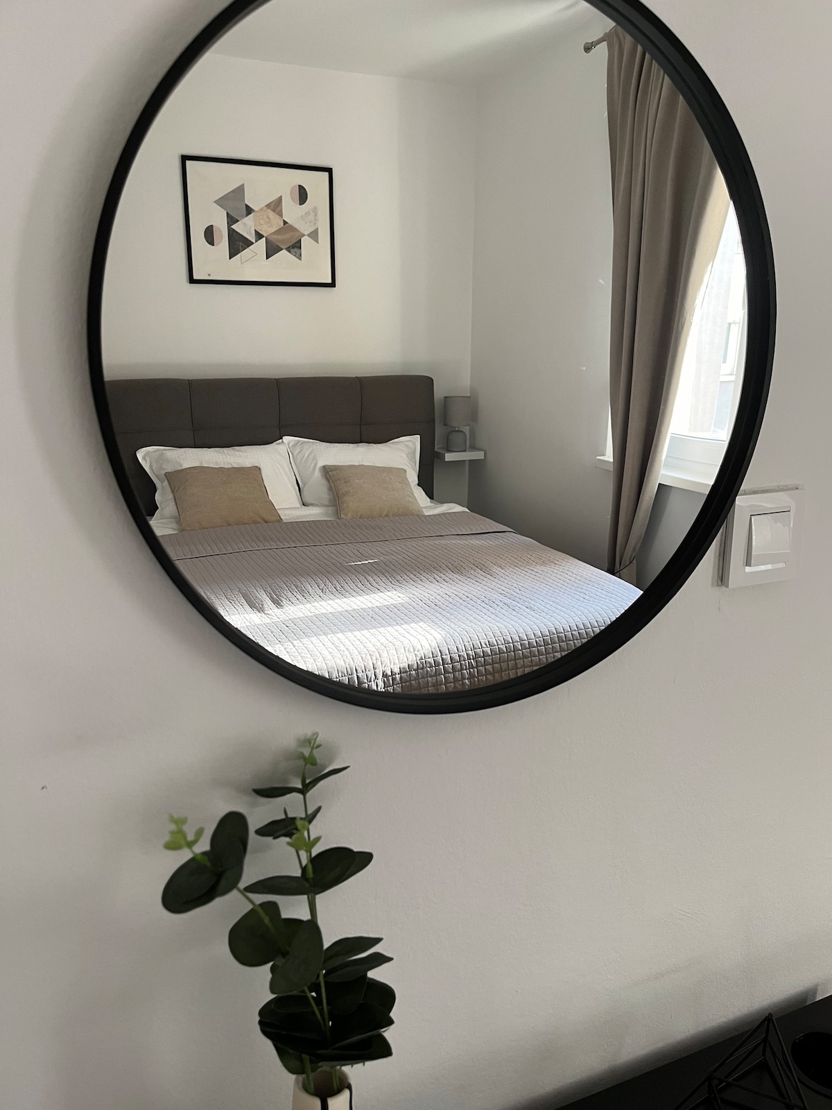 A round mirror reflects a cozy bedroom featuring a spacious bed with patterned pillows. A simple plant is positioned in the foreground, while soft curtains hang beside a window that allows natural light to enhance the room's ambiance.