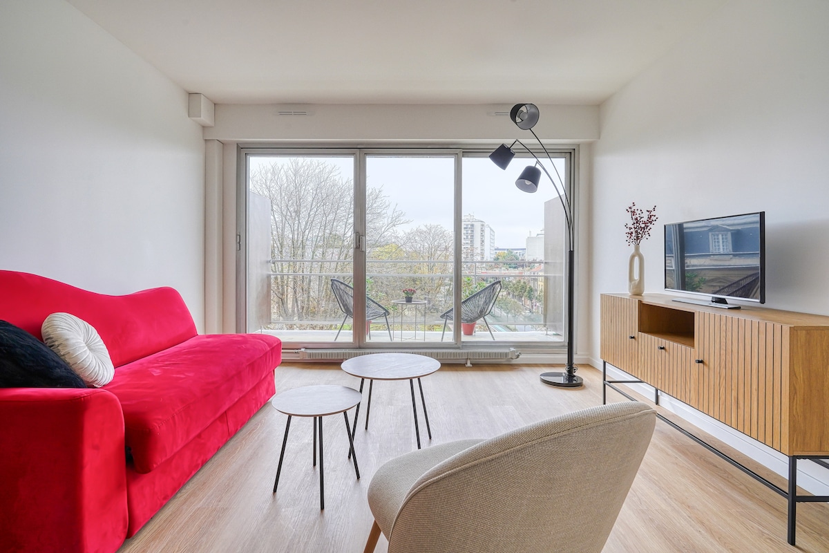The living area features a bright red sofa and two coffee tables set on a light wooden floor. A modern floor lamp provides additional lighting. Large glass doors lead to the balcony, revealing views of the surrounding area and greenery.