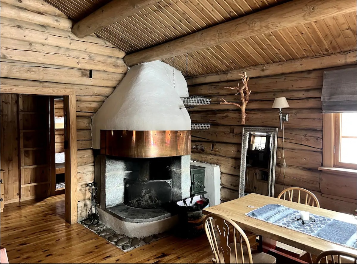 The living area features a large stone fireplace with a copper chimney, providing a rustic focal point against the log walls. A wooden dining table is positioned nearby, surrounded by light-colored chairs. Natural light filters through a window adorned with simple grey curtains.