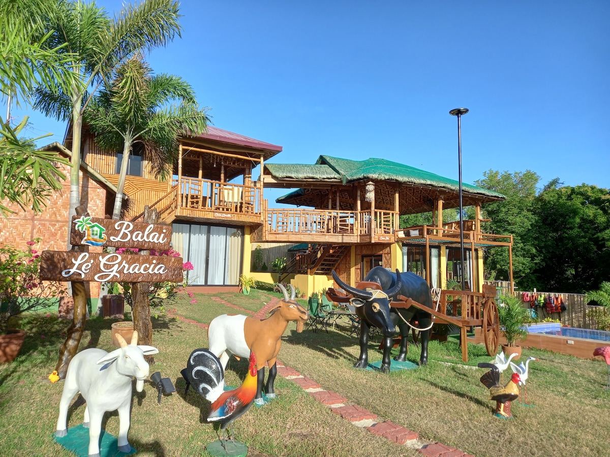 A charming farmhouse is depicted with a two-story wooden structure featuring a vibrant green roof. The landscaped garden showcases colorful animal sculptures and tropical plants, while a wooden sign labeled 'Balai Le Gracia' is prominently displayed in the foreground.