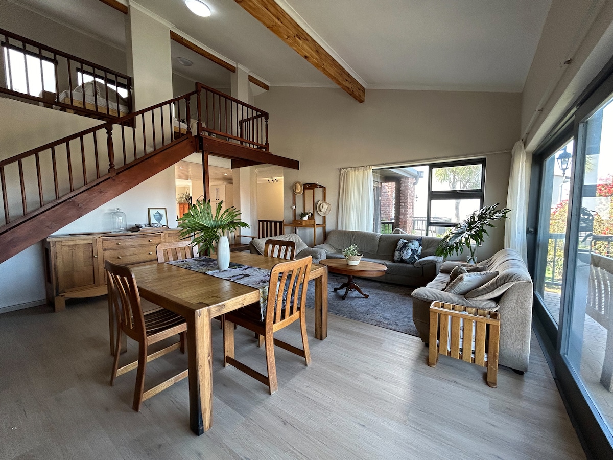 An open living and dining area features a wooden dining table surrounded by four chairs. A cozy seating arrangement with a sofa and side chairs is positioned near large windows, allowing natural light to fill the space. Exposed wooden beams add an architectural detail.