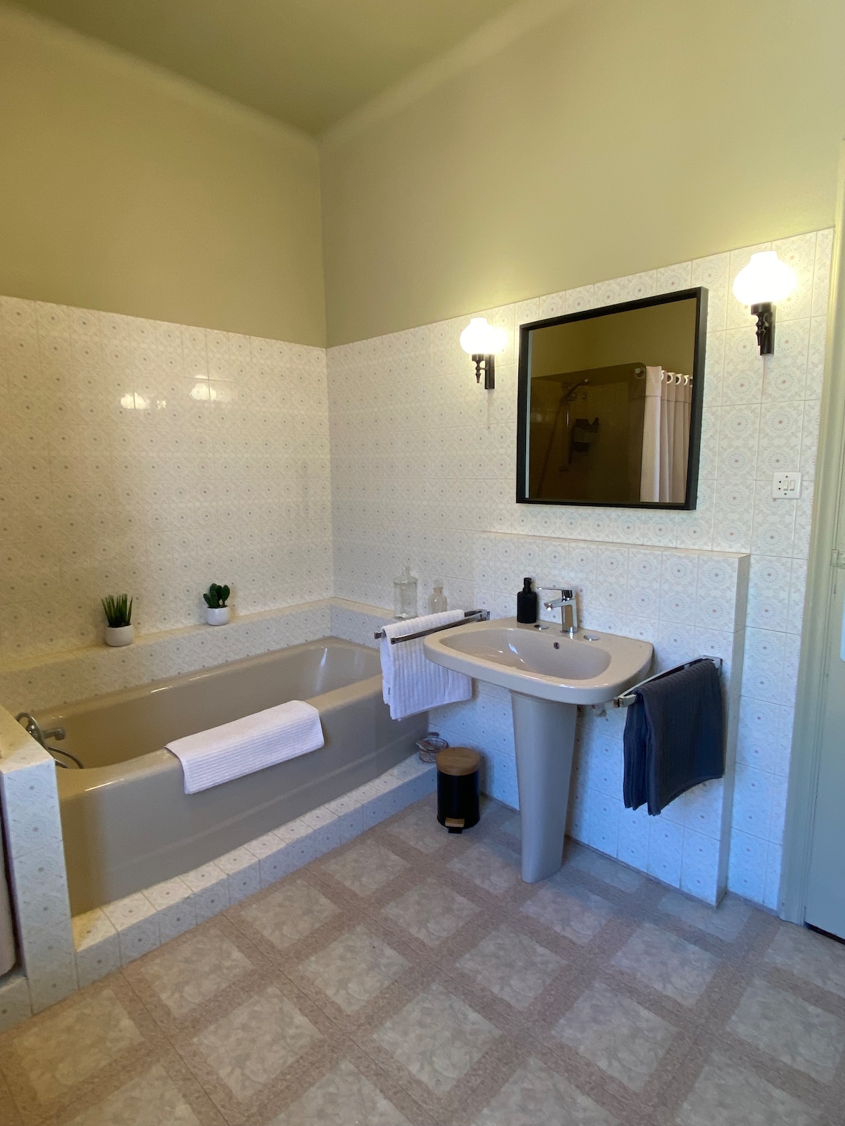 The bathroom features a spacious tub alongside a sink with a modern faucet. Elegant wall sconces provide soft lighting. A dark-framed mirror hangs above the sink, and decorative elements include a small plant and neatly arranged towels. Floor tiles exhibit a subtle pattern.