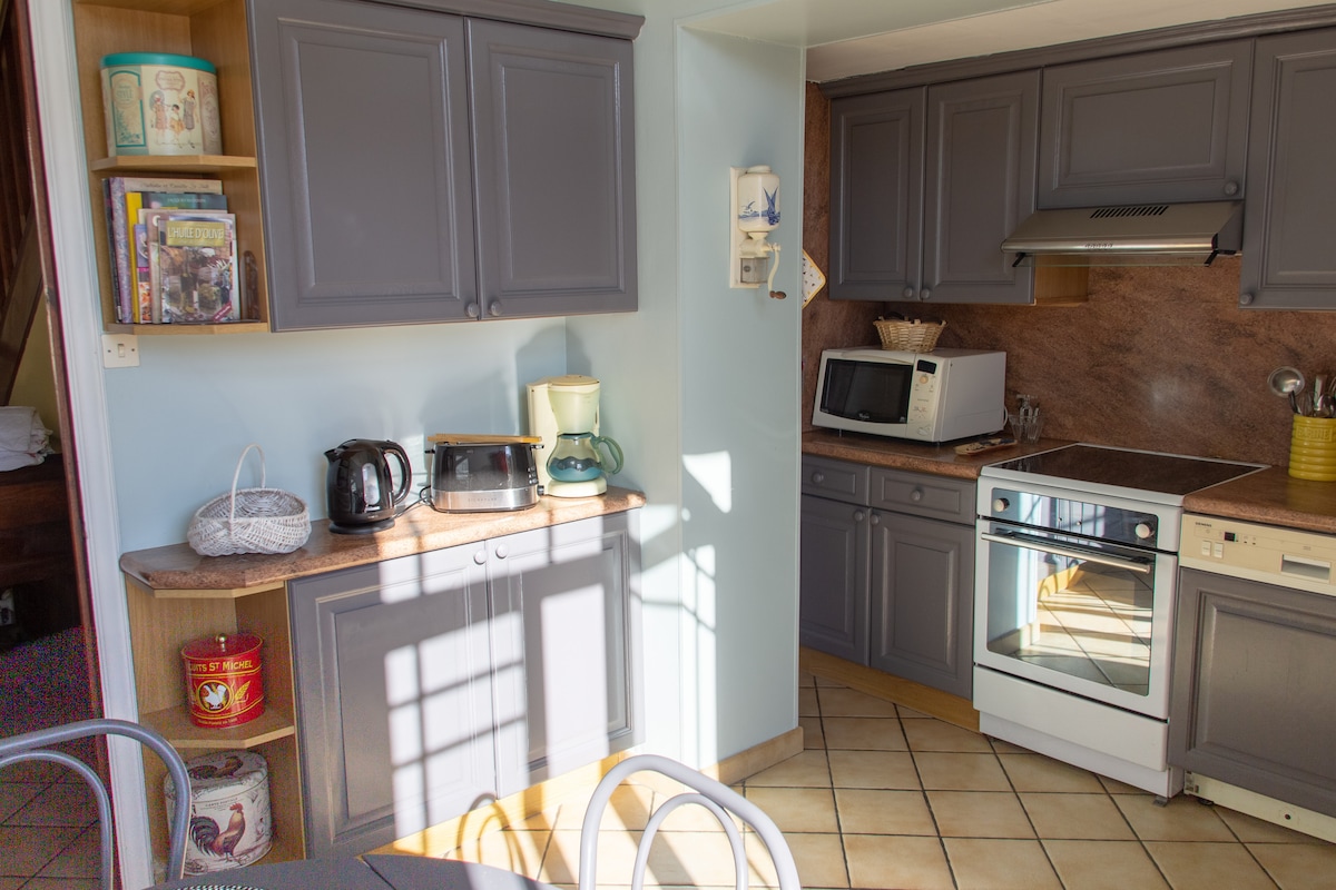 A well-equipped kitchen featuring dark cabinetry and a spacious countertop. Appliances include a microwave, coffee maker, and kettle, arranged alongside a sink. Natural light illuminates the room, highlighting the tiled floor and nearby dining area with a round table and chairs.