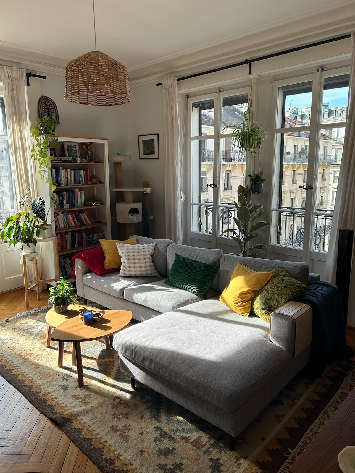 A cozy living area features a sectional sofa adorned with colorful cushions. Natural light streams through large windows, illuminating a small wooden table and a bookshelf filled with books. Houseplants add a touch of greenery to the warm space.
