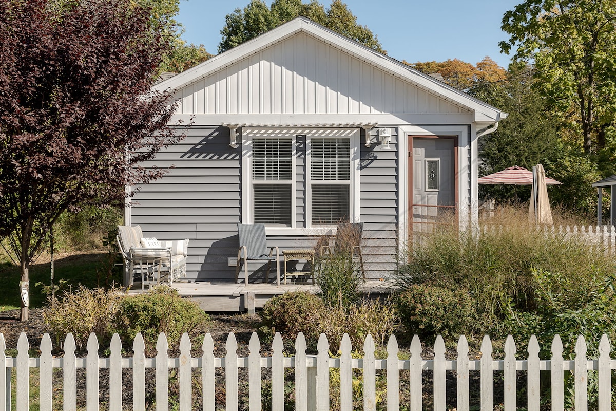 A charming gray cottage is surrounded by lush greenery, with a welcoming front porch furnished with comfortable seating. A white picket fence encloses the property, providing a quaint touch to the landscape. The entrance is framed by large windows, allowing natural light to flow inside.