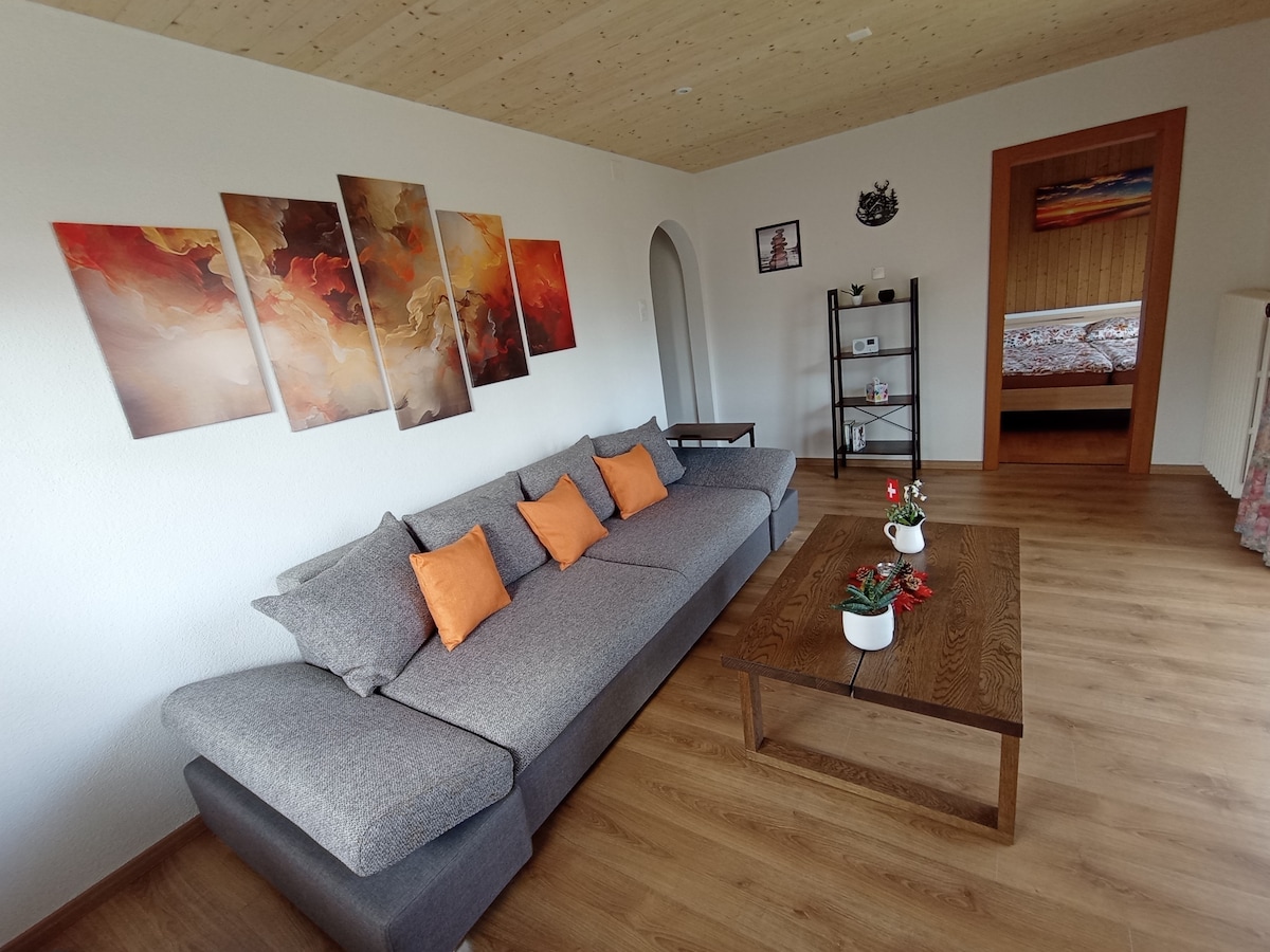 A spacious living area features a large, grey couch adorned with orange cushions. A wooden coffee table rests in front, accompanied by decorative plants. Colorful artwork adorns the wall, while a shelf houses decorative items. A doorway leads to another room, suggesting a cozy layout.