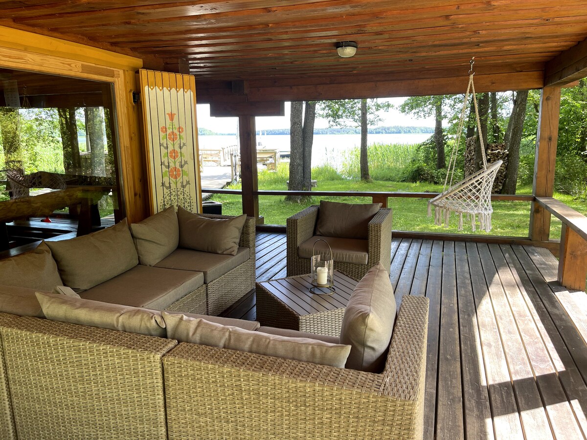 A covered outdoor seating area features a comfortable arrangement of wicker furniture, including a sectional sofa and a coffee table. A hammock chair hangs nearby, providing a relaxing spot. Tall trees and a scenic lake view create a peaceful backdrop.