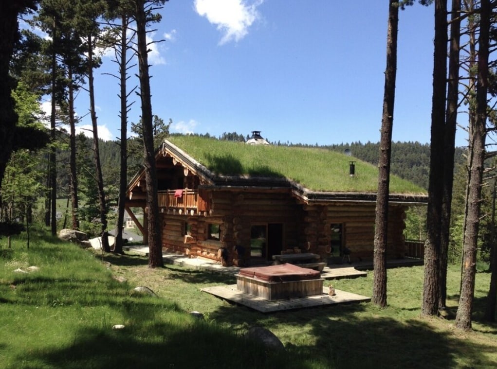 A wooden cabin with a sloping grass roof is nestled among tall trees. A small outdoor hot tub is placed on a stone patio, offering a natural setting. Scenic mountain views are visible in the background, enhancing the tranquil environment.