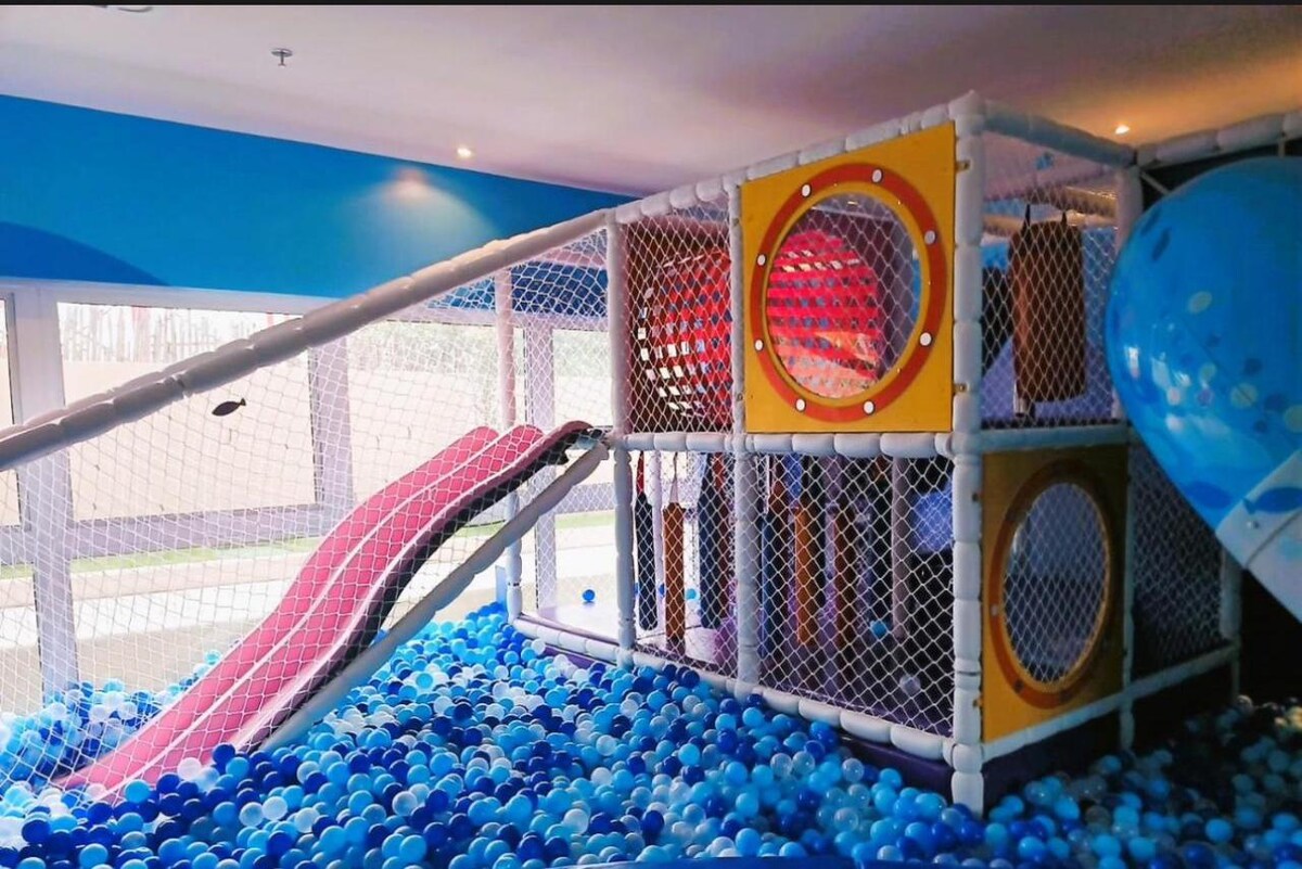 A colorful children's play area features slides and a netted play structure surrounded by a sea of blue and white balls. Large windows allow natural light to illuminate the space, creating an engaging environment for kids.
