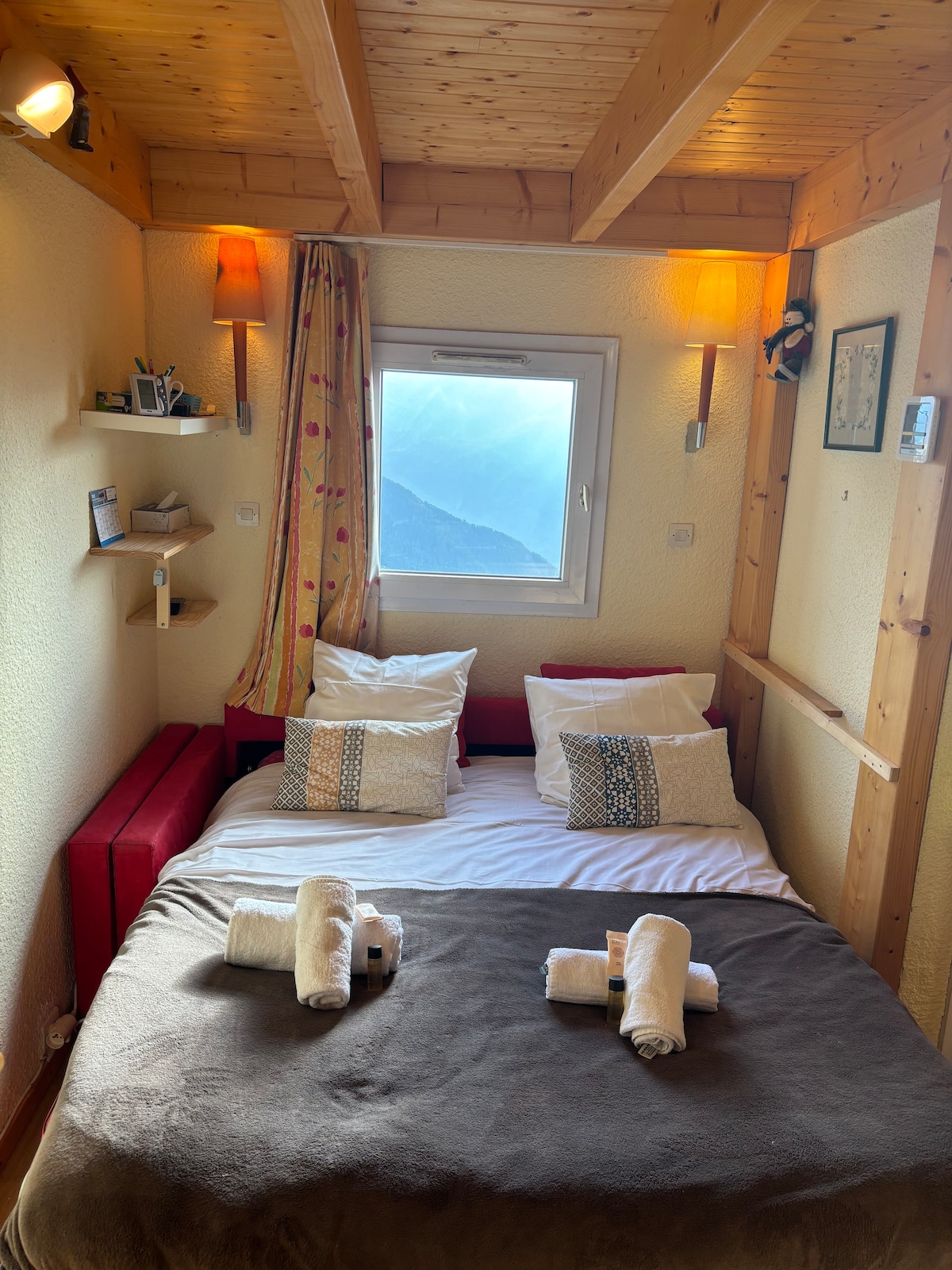 A cozy bedroom is featured, showcasing a spacious bed covered with a brown blanket. The bed is adorned with two pillows and neatly arranged towels. Wooden beams are visible above, and a window allows natural light to fill the room, providing a view of the mountains.