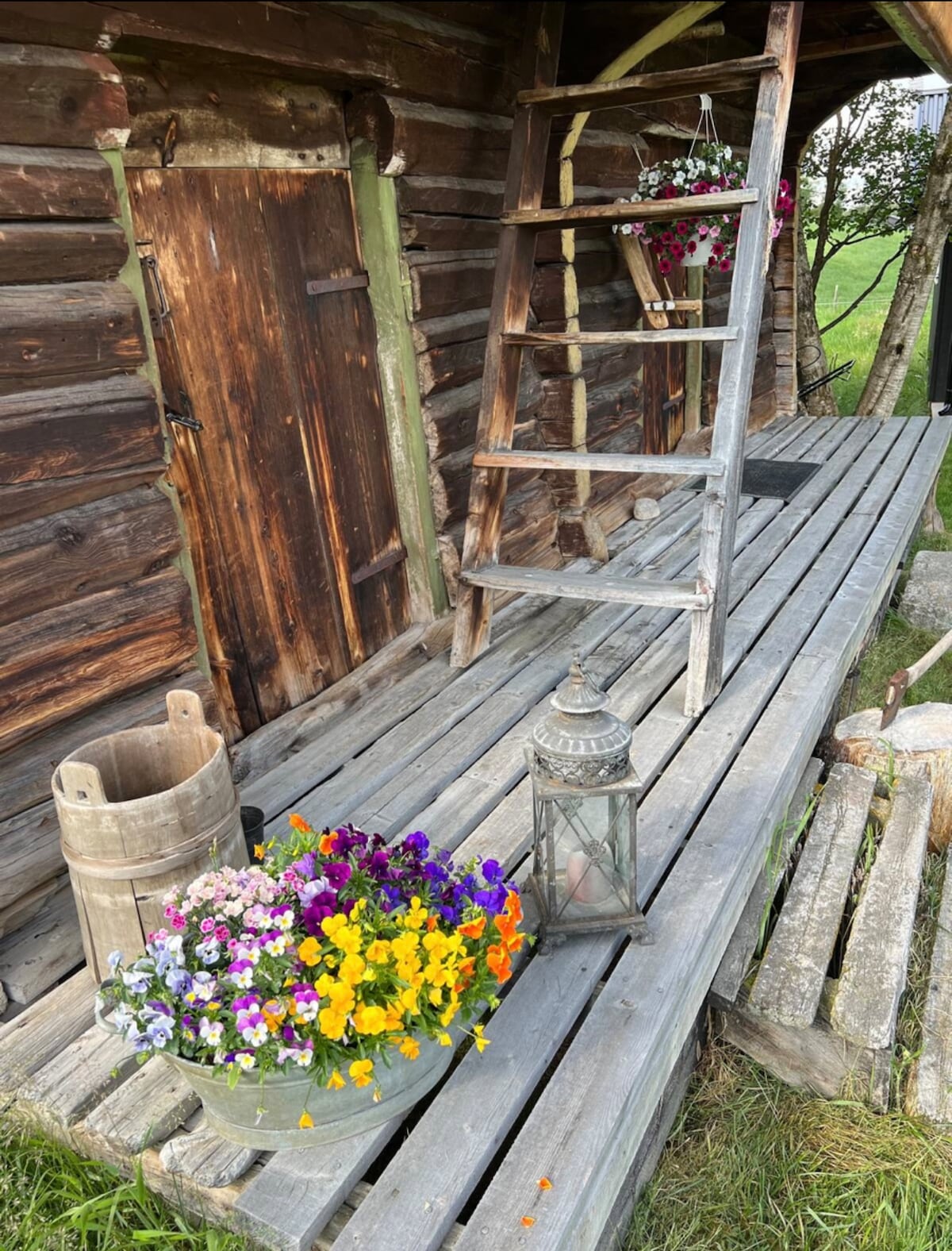 A wooden porch features a rustic ladder, flower pots with colorful blooms, and a lantern. The weathered wood adds character, while a wooden barrel rests nearby, enhancing the natural setting.