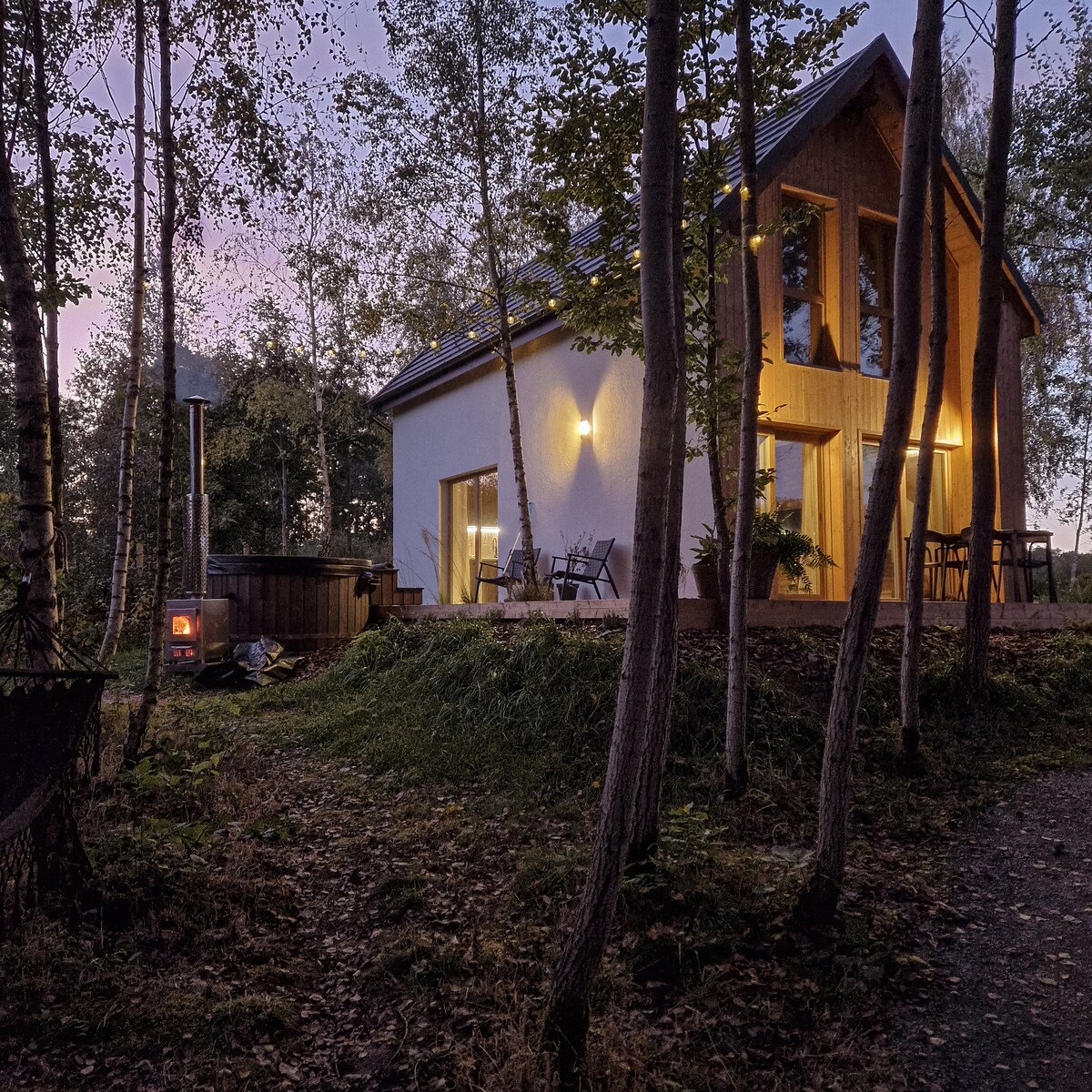A modern cabin is nestled among trees, featuring large windows that illuminate the interior. A private hot tub is visible on the patio, with seating arranged around a natural setting. A sauna and a wood-burning stove can be seen nearby, enhancing the peaceful surroundings.