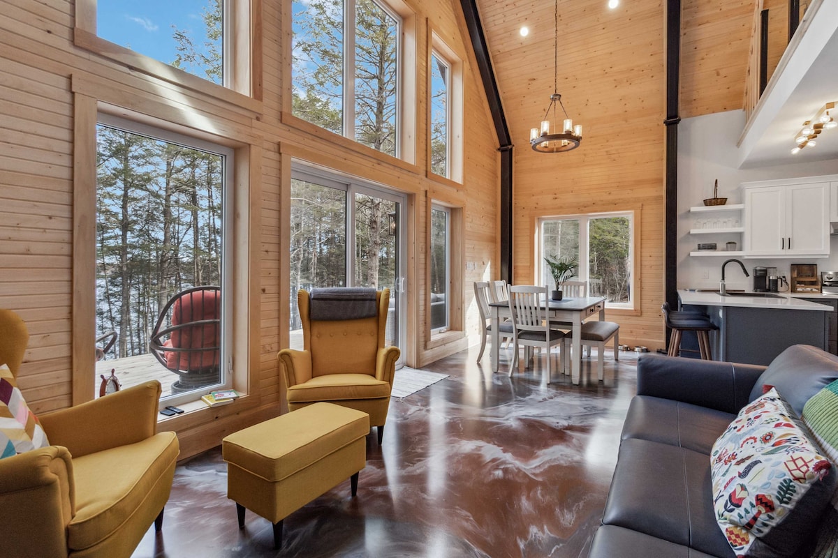 A spacious living area features tall windows allowing natural light to fill the space, showcasing views of trees and the nearby lake. Natural wood walls and ceiling add warmth, while modern furnishings, including a couch, armchairs, and a dining table, create a comfortable gathering place.