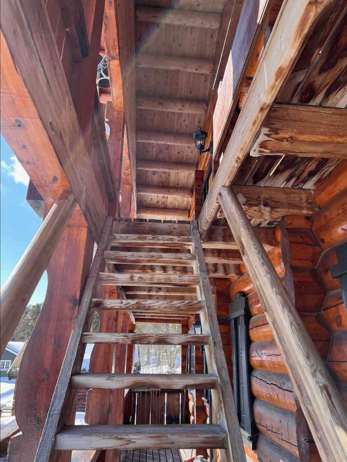 A wooden staircase leads to the second floor of a log building. The sturdy steps, crafted from timber, reflect the natural finish of the logs surrounding them. Soft sunlight filters through, casting gentle shadows along the structure. The outdoor ambiance suggests a serene, rustic setting.
