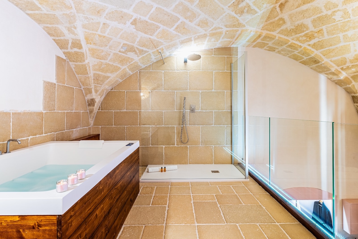 A luxurious bathroom features an inviting whirlpool tub alongside a modern walk-in shower. The stone arch ceiling complements the natural stone walls, while warm lighting creates a soothing ambiance. Light-colored tiles extend across the floor, enhancing the spacious feel of the area.