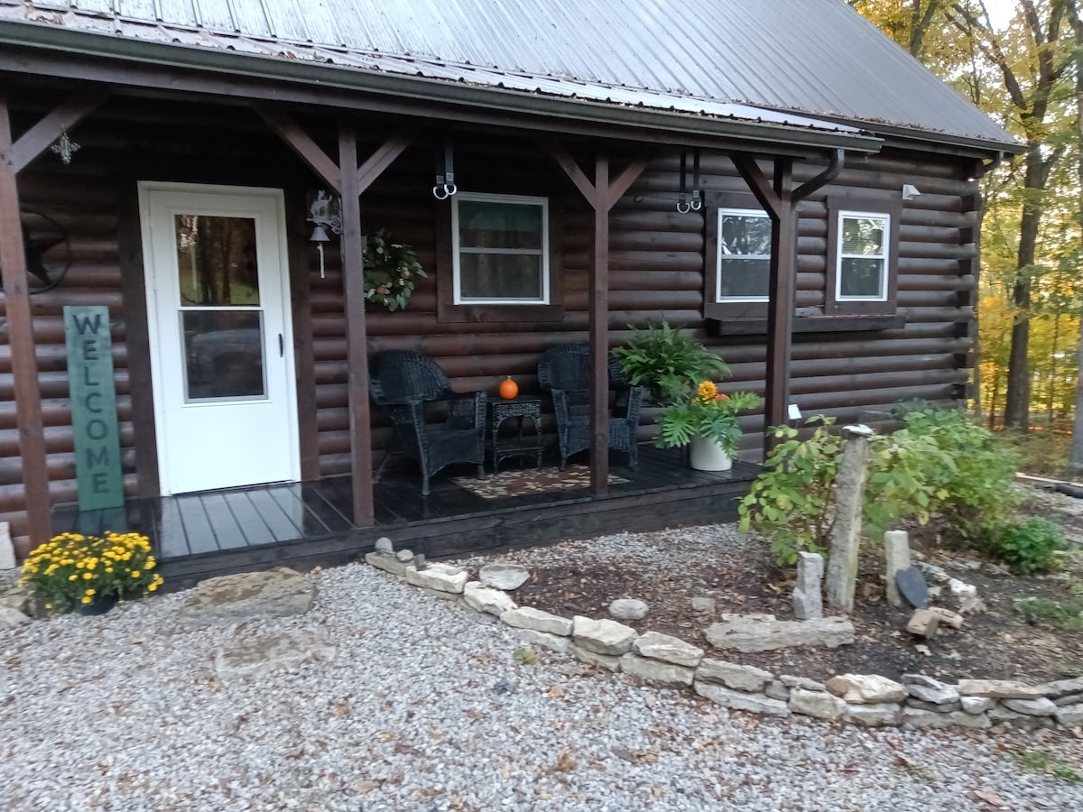 The entrance of the cabin features a wooden porch with two dark wicker chairs and a small table. A welcome sign is prominently displayed next to the door, while vibrant yellow and orange flowers are placed in pots. A mix of gravel and stone outlines the garden area.