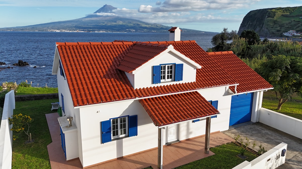 A charming cottage is set against the backdrop of Pico Island, featuring a red-tiled roof and blue window shutters. The property is surrounded by water, with a spacious outdoor area visible. Impressive cliffs rise in the distance, enhancing the coastal scenery.