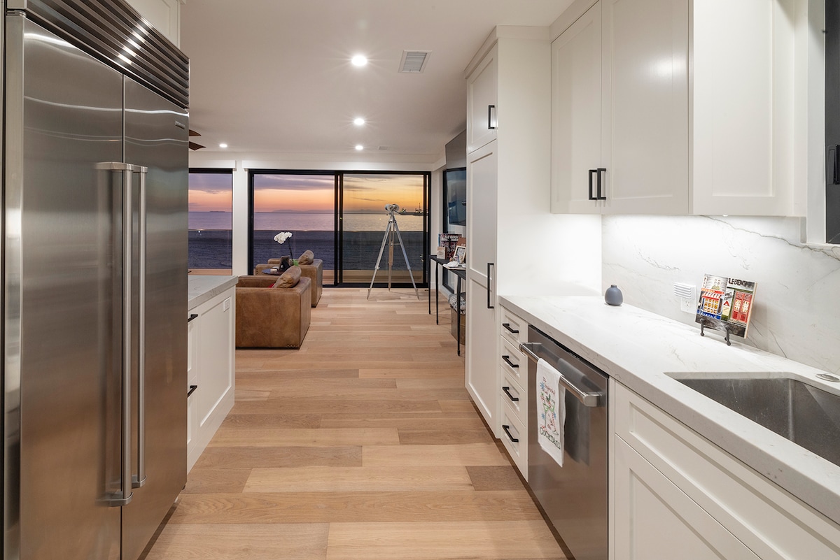A modern kitchen is showcased with stainless steel appliances, including a large refrigerator. The open layout allows for views of the ocean through expansive glass doors. Light wood flooring extends into a living area, featuring a comfortable sofa and a telescope, with sunset colors visible outside.