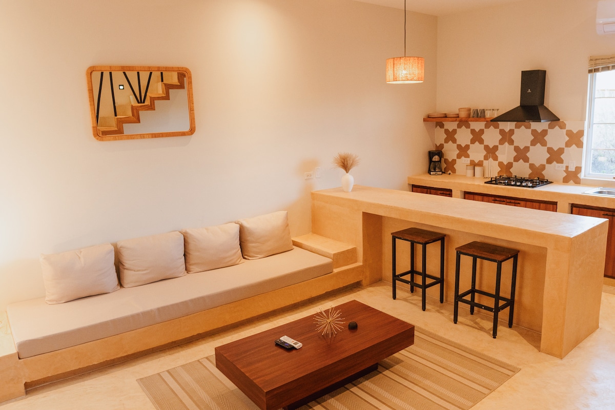 A living area is presented with a neutral color palette, featuring a low-slung couch and a wooden coffee table. Two bar stools are situated at a countertop, with patterned tiles visible in the background. Natural light fills the room from a nearby window.