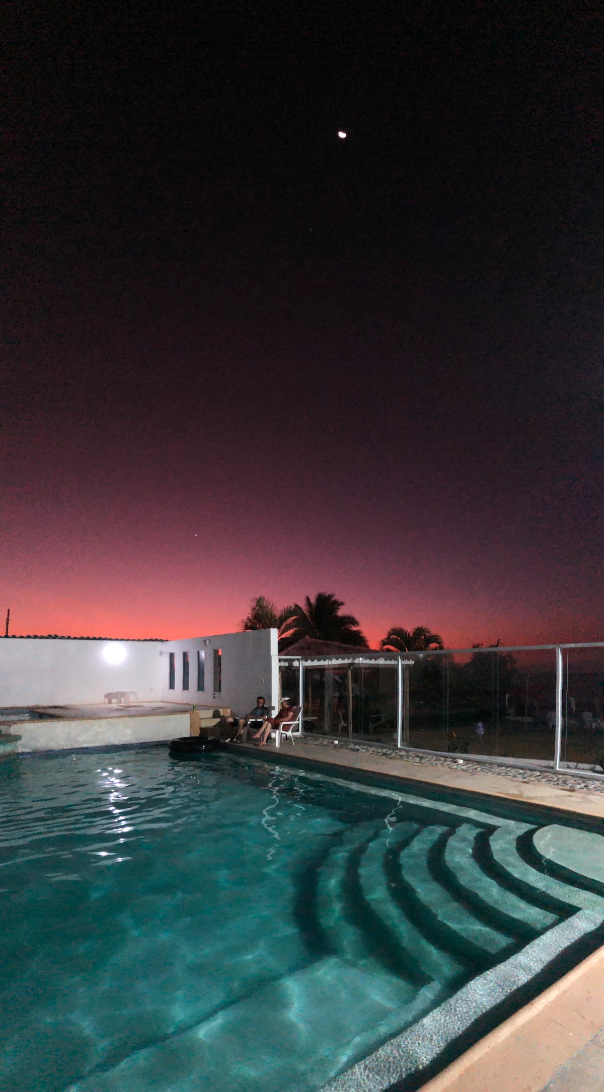 A tranquil pool reflects the gradient of a twilight sky transitioning from deep purple to soft pink. The pool's entrance features gentle steps, while lounge chairs are positioned nearby. Palm trees are visible in the background, silhouetted against the evening light.