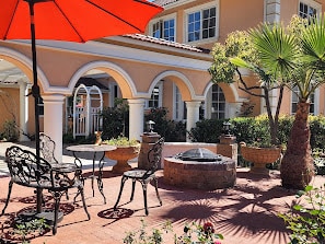 A charming outdoor patio area features iron wrought chairs surrounding a stone fire pit. An umbrella provides shade over one seating arrangement, while surrounding greenery and planters enhance the natural environment. The architecture showcases elegant arches and spacious windows in the backdrop.