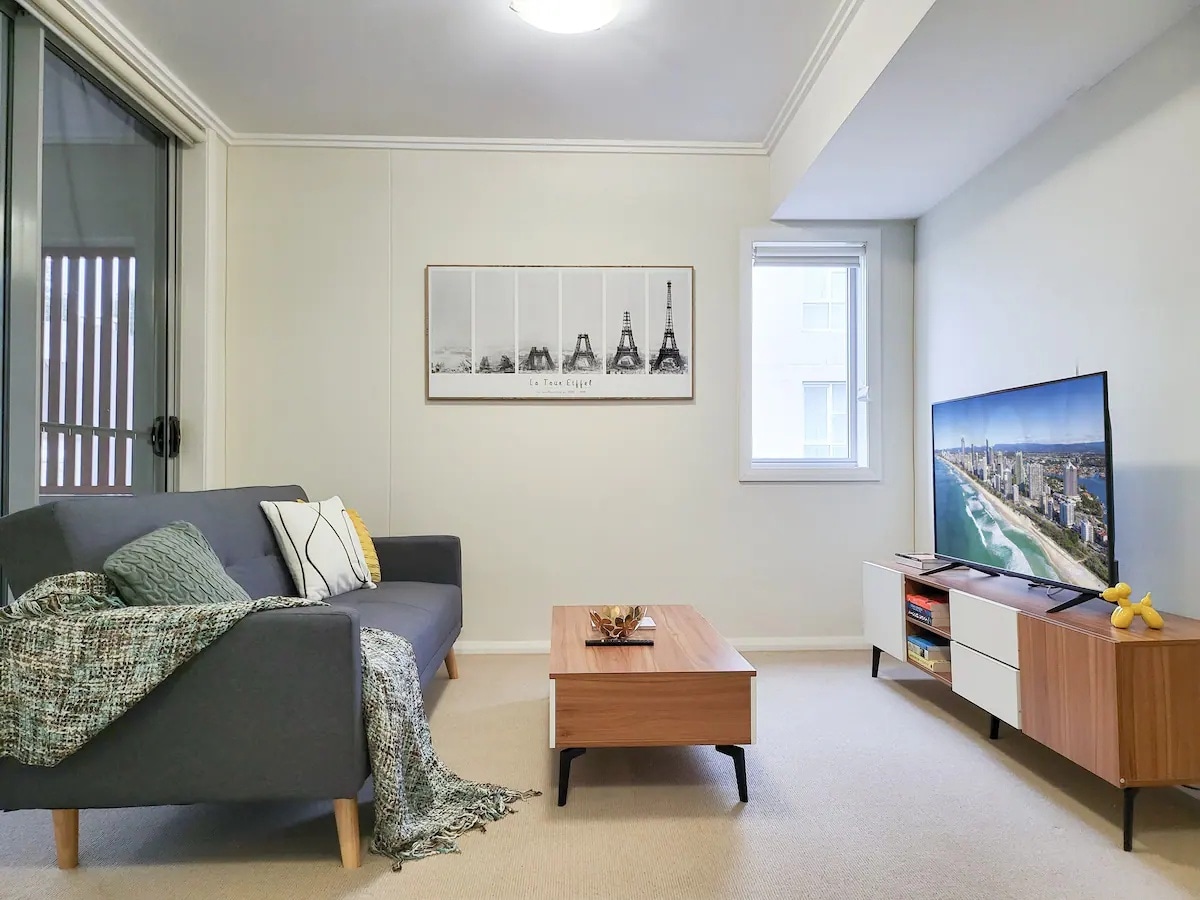 The living room features a comfortable gray sofa adorned with a textured throw, positioned near a wooden coffee table. A large flat-screen TV is mounted on a sideboard, with a wall art piece showcasing iconic landmarks. Natural light enters through the balcony doors.