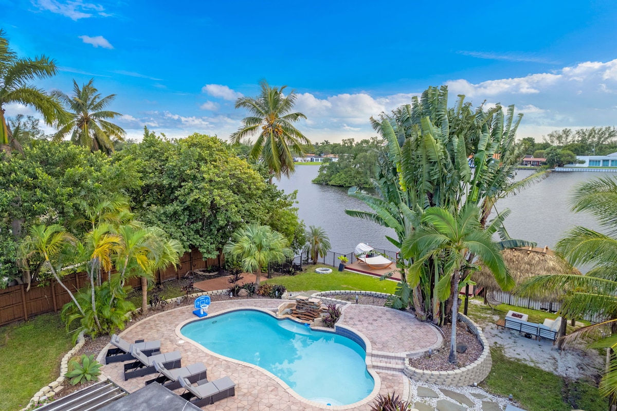 An aerial view captures a curved pool surrounded by lounge chairs, with lush tropical landscaping and palm trees. A tiki hut is visible nearby, beside a calm body of water that reflects the sky. The scenery provides a relaxed outdoor atmosphere.