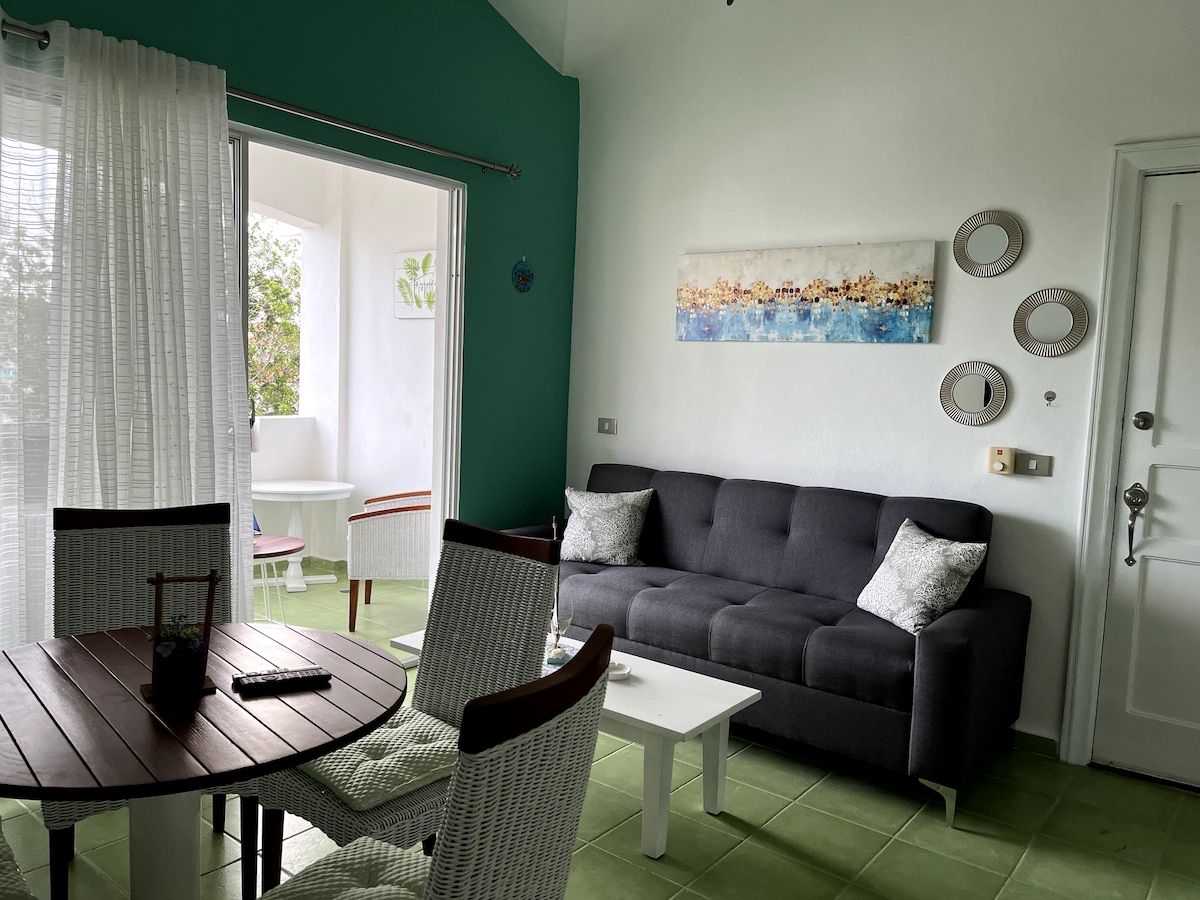A living area features a dark gray sofa with decorative cushions, accompanied by a round dining table and four white chairs. Light streams through sheer curtains, illuminating the green tiled floor. A wall art piece with abstract designs is displayed above the sofa.