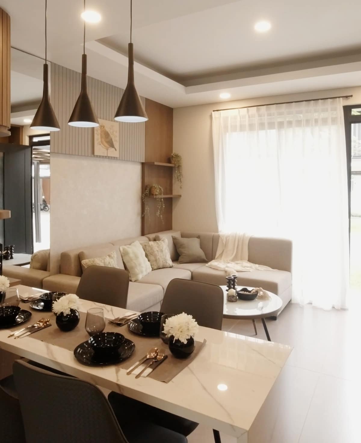A modern dining area features a sleek table set for meals, complemented by black dishware and floral centerpieces. Soft lighting is provided by pendant fixtures above the table. A cozy corner sofa offers a relaxed seating option, with large windows allowing natural light to fill the space.
