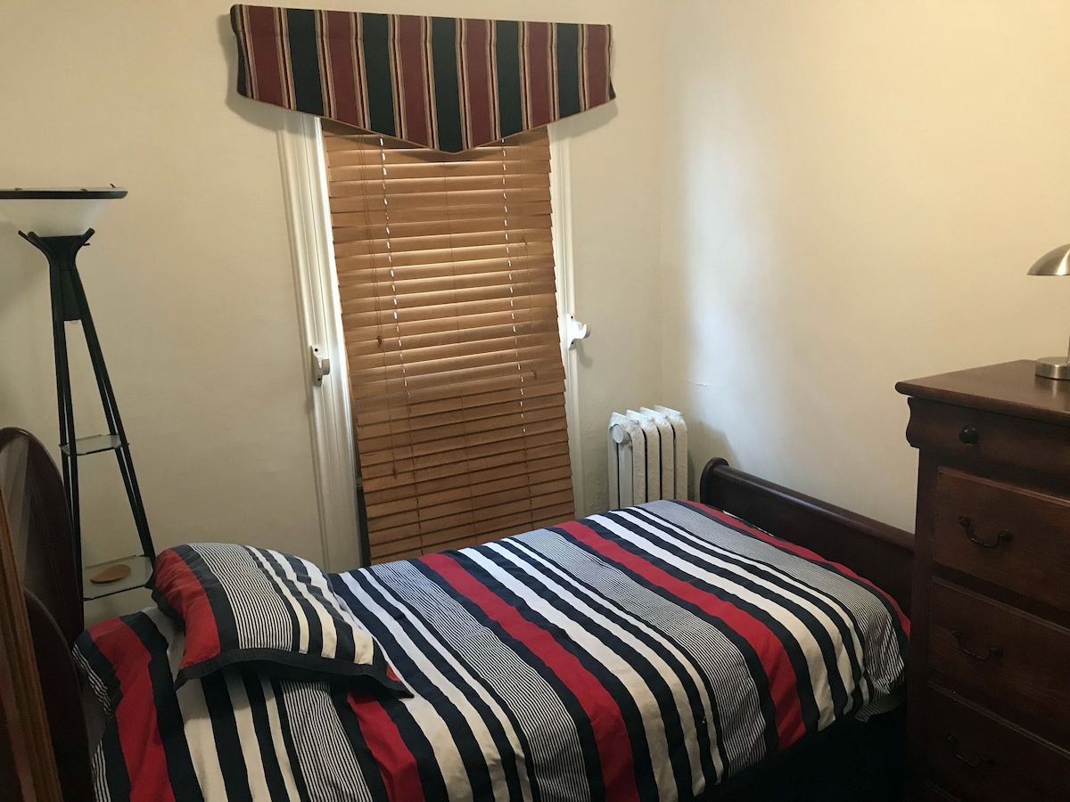 A well-arranged cozy room features a twin bed with striped bedding next to a wooden dresser. A reading lamp is positioned on the dresser. Blockout blinds are drawn over a window, allowing for light control, while a radiator is present for warmth.