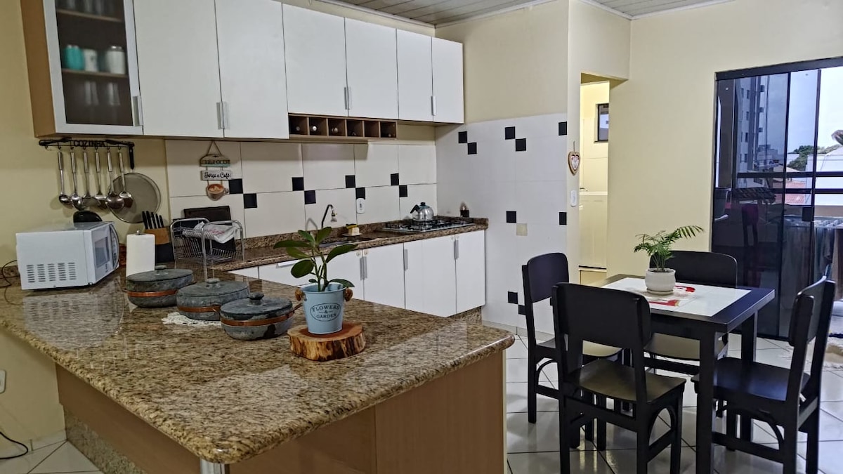 The kitchen area features modern cabinetry and a spacious granite countertop. Cooking essentials are provided, including a microwave. A dining table for four is positioned nearby, along with a small potted plant. The brightly lit space opens to a balcony area.