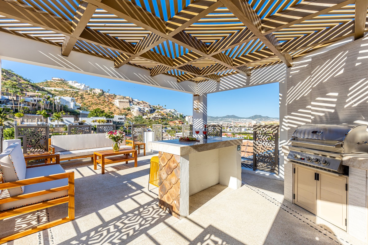 An outdoor terrace is featured, showcasing a shaded seating area with wooden furniture and a BBQ grill. The backdrop includes scenic hills, while natural light filters through the decorative overhead structure, creating a pattern on the surface below. Fresh flowers are arranged on the counter.