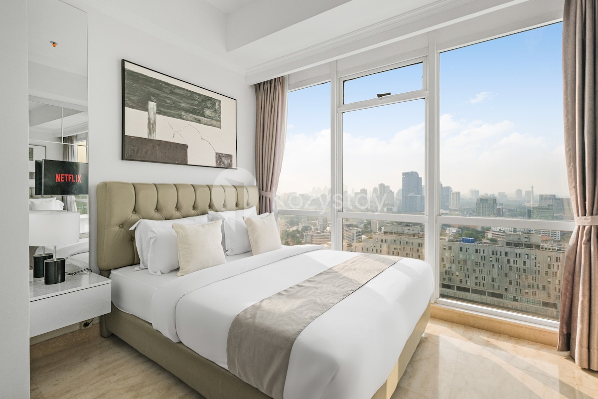 A modern bedroom features a queen size bed with crisp linens and decorative pillows. Large windows provide expansive city views, while a stylish painting adorns the wall. A flat-screen TV is mounted within view, ensuring entertainment options are easily accessible.