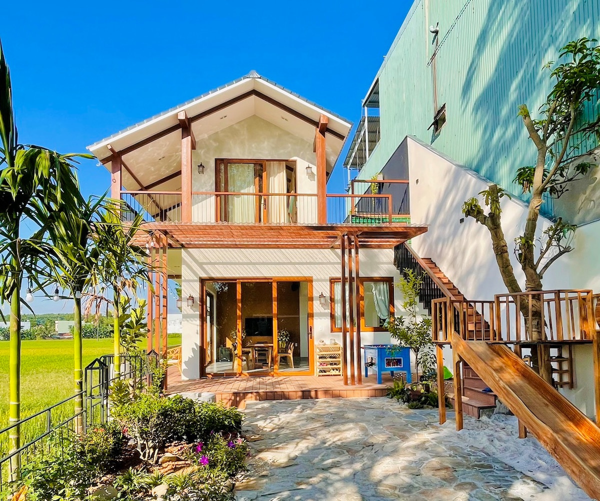 A two-story house with a spacious balcony overlooks a vibrant green field. The exterior features a combination of light and dark wood accents, framed by lush greenery and a stone pathway leading to the entrance. Sunlight bathes the structure, highlighting its inviting design.