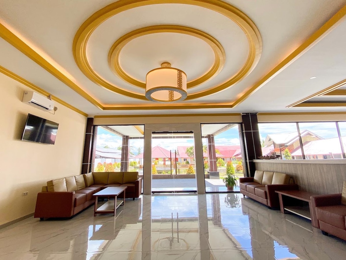 A spacious lobby area is showcased, featuring a stylish ceiling design with a round light fixture. Comfortable brown leather seating is positioned to encourage conversation, with large windows allowing natural light to fill the space. The well-maintained floor reflects the airy atmosphere.