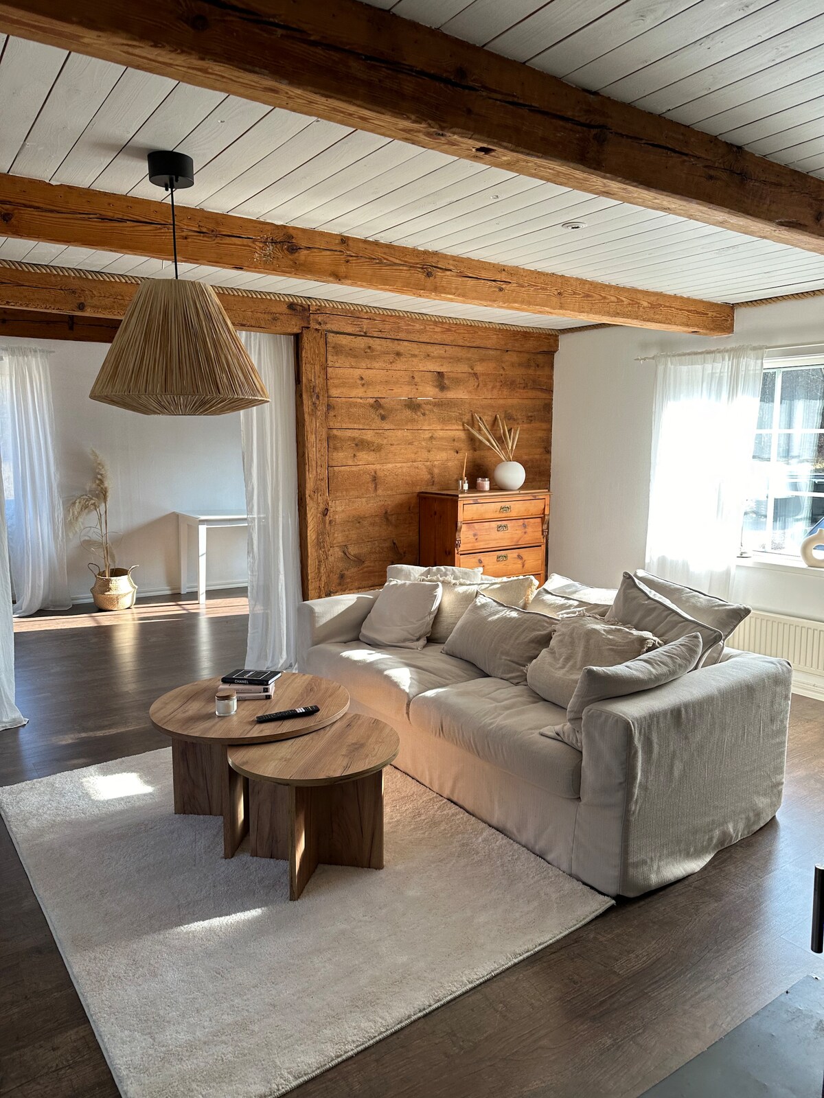 A living room features natural wood beams and a light color palette. A cozy sofa is situated around a circular coffee table. Soft textiles and decorative touches create a relaxed ambiance, with abundant natural light streaming through the large window dressed in sheer curtains.
