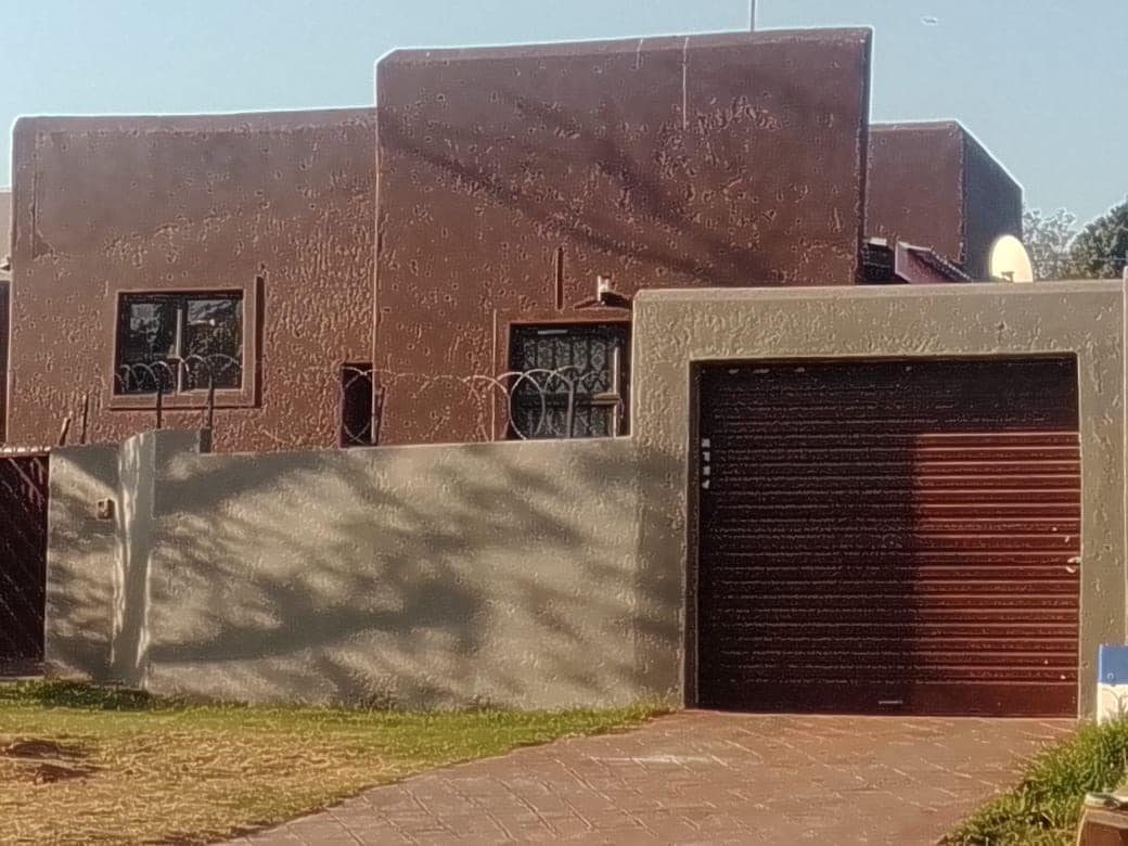 The exterior of a three-bedroom property is shown, featuring a unique two-tone facade with brown walls and a grey accent. A garage door is visible, alongside a wall topped with wire fencing. The front yard consists of a well-maintained lawn, edged by a paved path.