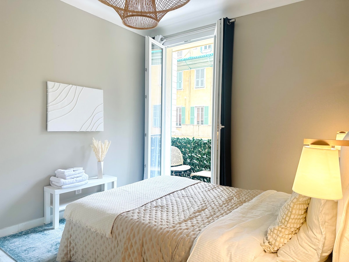 The bedroom features a spacious bed covered with a neutral-toned quilt and accent pillows. A sliding glass door opens to a private balcony, while a decorative light fixture hangs from the ceiling. A small table is placed beside the bed, with neatly arranged towels displayed.