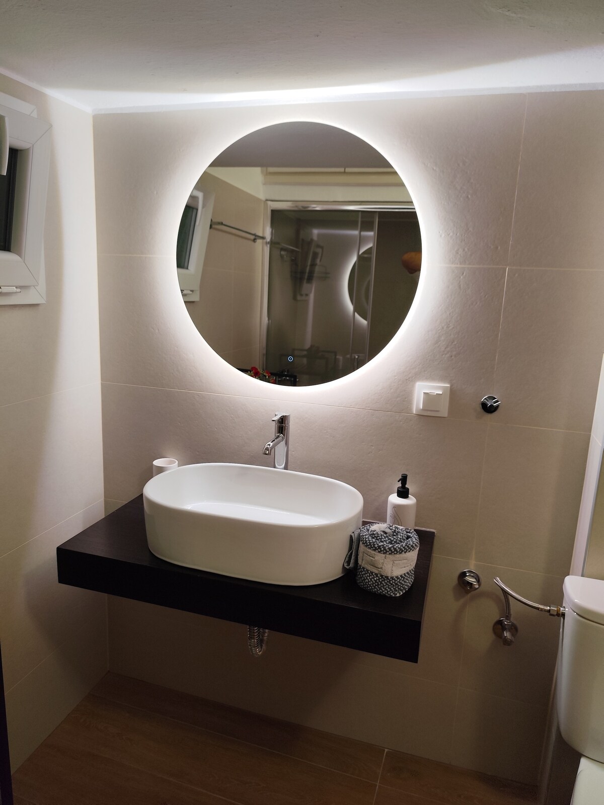 A modern bathroom is highlighted by a round mirror with integrated lighting above a sleek, rectangular sink. A dark wooden shelf holds toiletries, complemented by a small decorative item. Subtle tiles and a shower enclosure add to the overall clean and functional design.