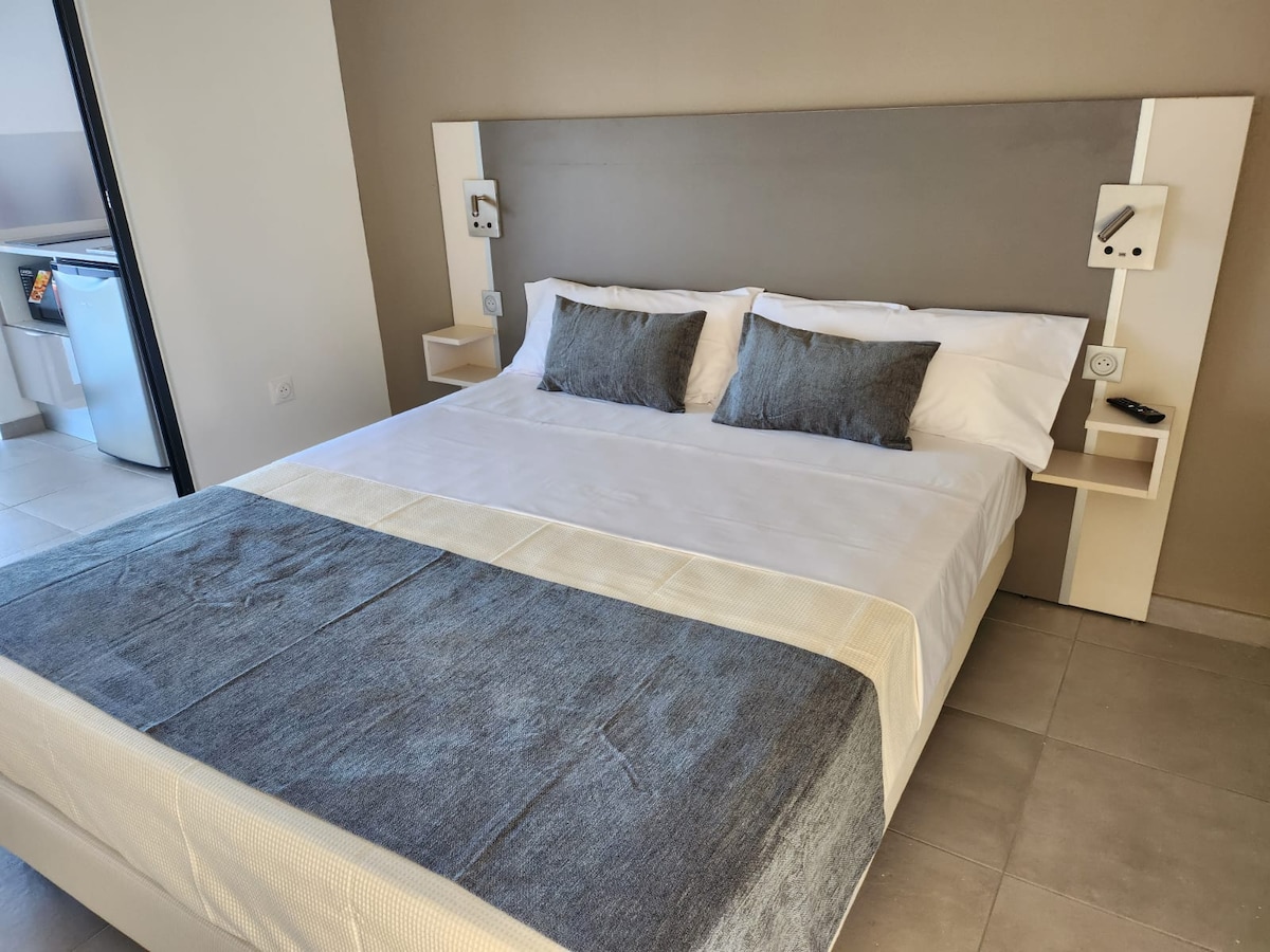A spacious bed is centered against a neutral-colored wall, flanked by two minimalist nightstands. Soft gray pillows and a textured blanket add a touch of comfort. In the background, a kitchenette is partially visible, providing convenient amenities.